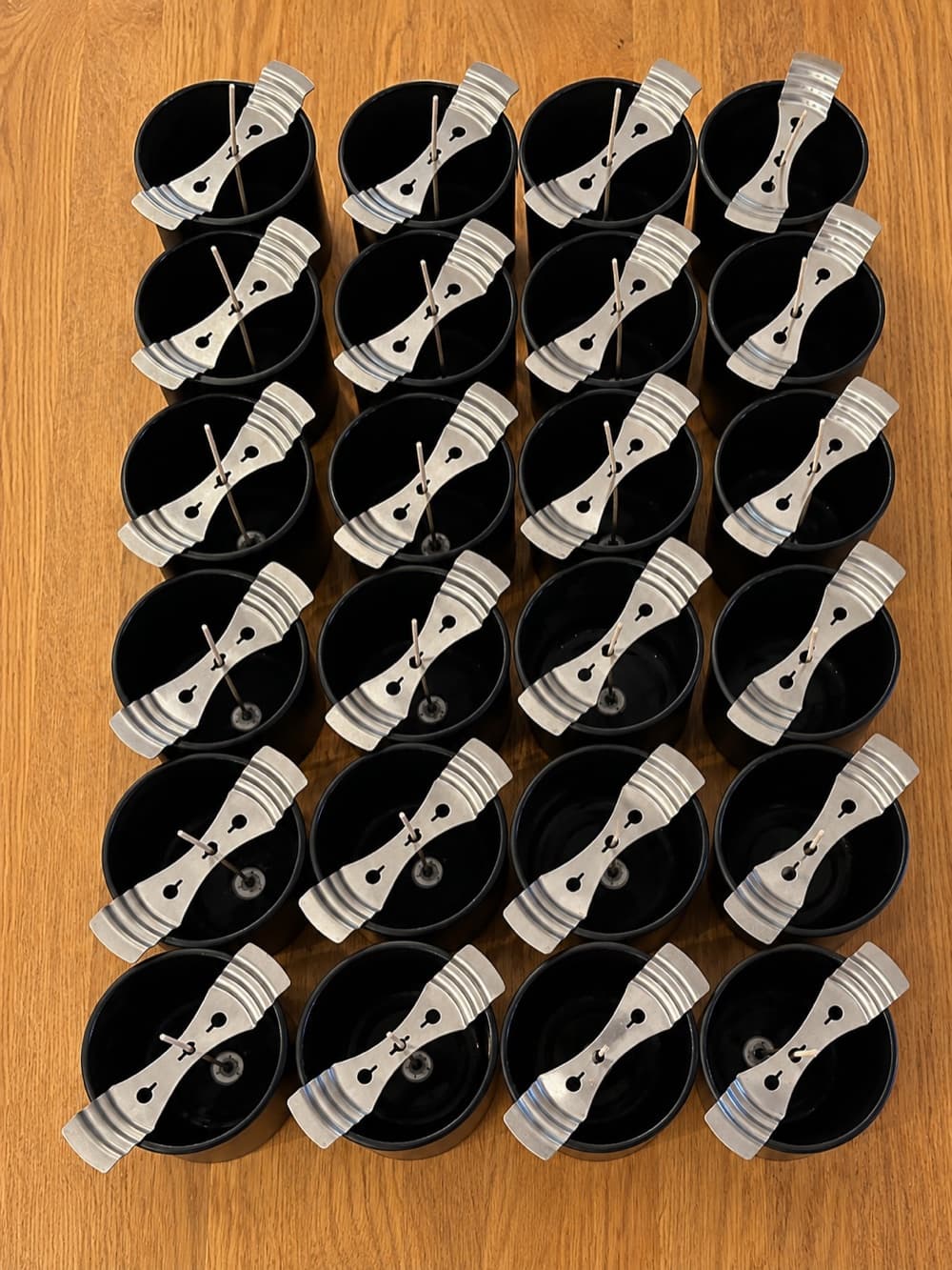 Two dozen empty black jars laid out on a wooden table, each with its wick held upright by a metal centring bar.